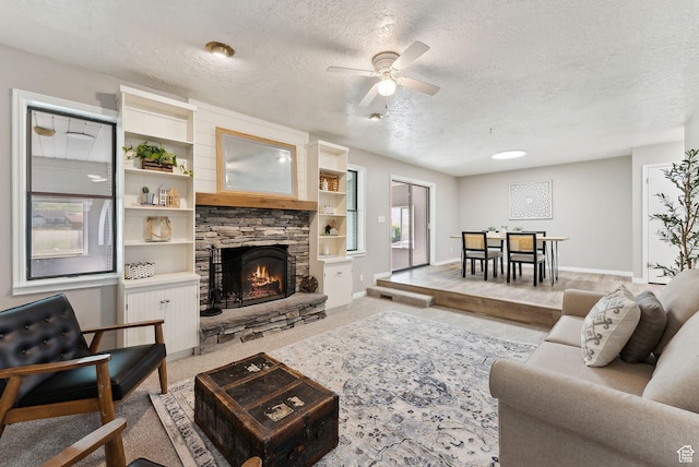 living room with ceiling fan, a stone fireplace, a textured ceiling, baseboards, and light colored carpet