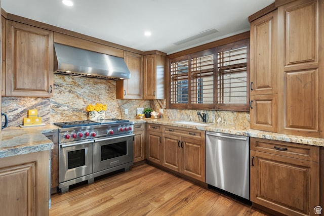 kitchen featuring appliances with stainless steel finishes, wall chimney range hood, a sink, backsplash, and light wood-style floors