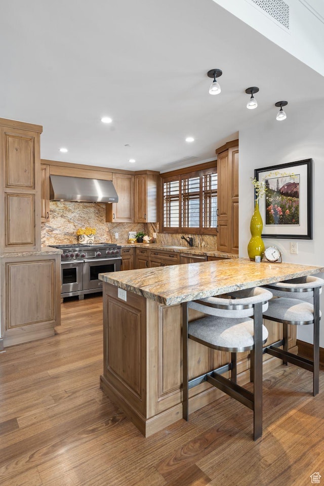 kitchen featuring wall chimney exhaust hood, stainless steel appliances, tasteful backsplash, light wood-style floors, and a kitchen bar