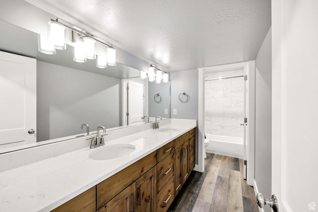 bathroom featuring wood finished floors, a textured ceiling, double vanity, toilet, and tub / shower combination