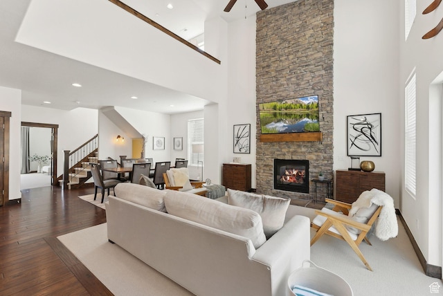 living area with ceiling fan, a towering ceiling, a stone fireplace, dark wood-style floors, and stairs