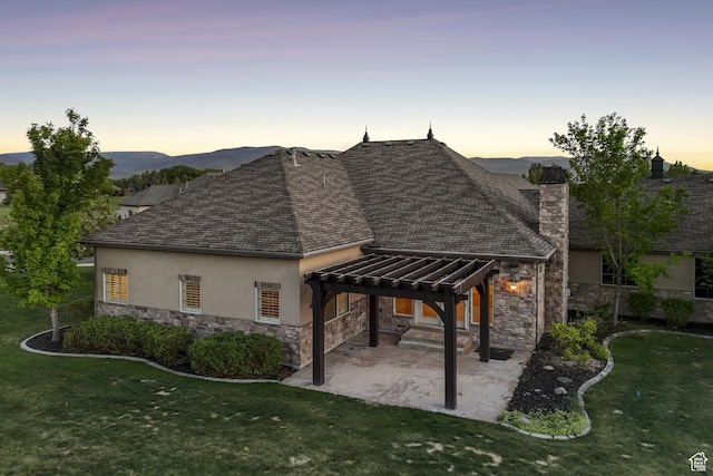 back of house at dusk featuring a pergola, stone siding, stucco siding, a patio, and a yard