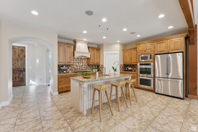 kitchen with stainless steel appliances, custom exhaust hood, arched walkways, light stone counters, and a sink