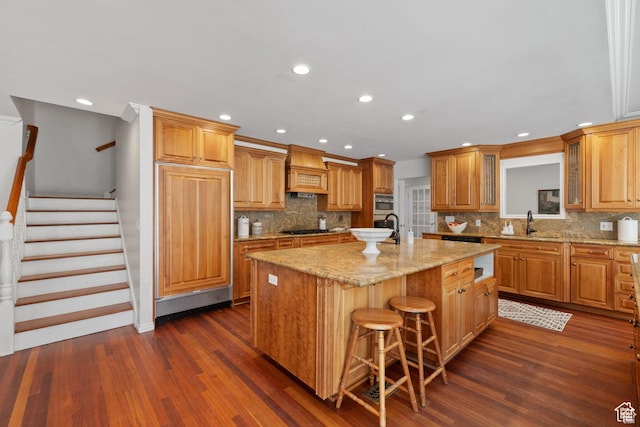kitchen with an island with sink, black cooktop, dark wood-style flooring, custom exhaust hood, and paneled built in fridge
