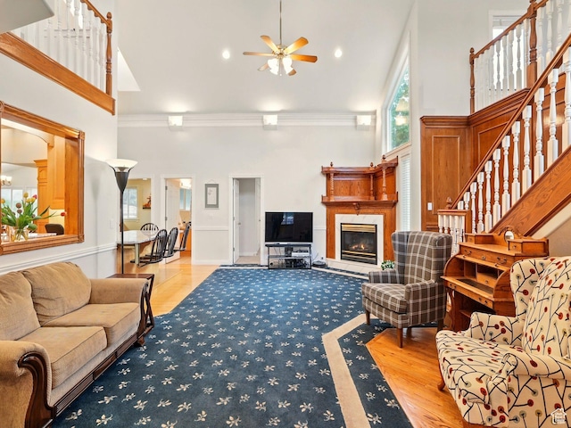 living room with ceiling fan, ornamental molding, a towering ceiling, a warm lit fireplace, and stairway
