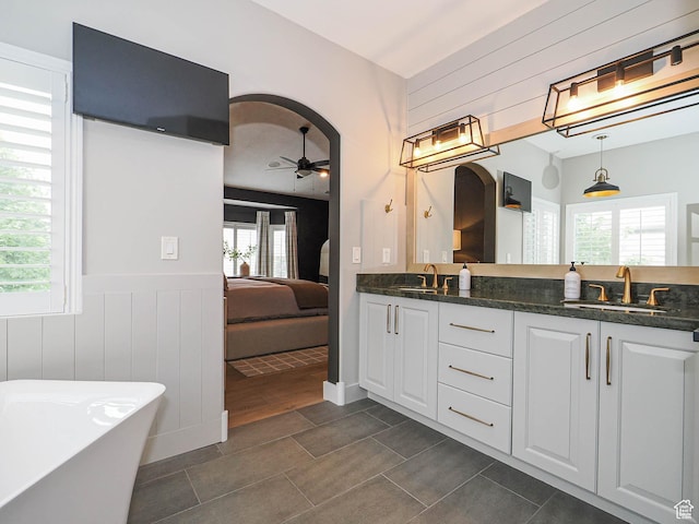 ensuite bathroom with ceiling fan, plenty of natural light, double vanity, a soaking tub, and wainscoting
