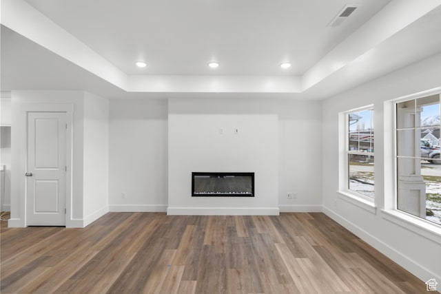 unfurnished living room featuring a raised ceiling, wood finished floors, a glass covered fireplace, baseboards, and recessed lighting