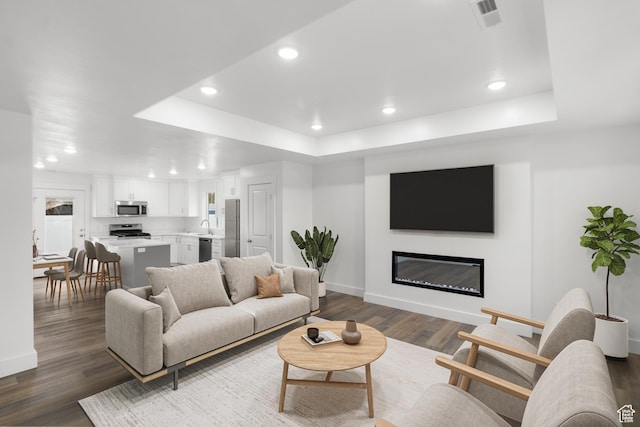 living room featuring a tray ceiling, baseboards, recessed lighting, dark wood-type flooring, and a glass covered fireplace
