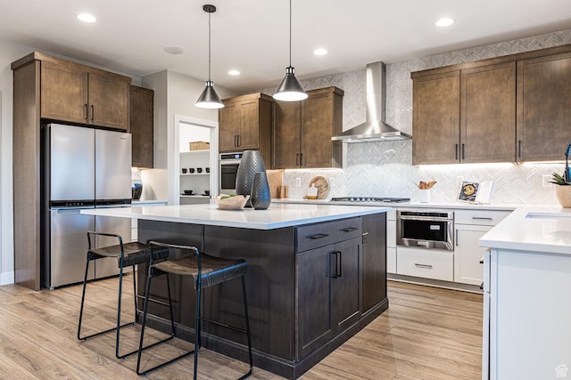 kitchen featuring appliances with stainless steel finishes, wall chimney range hood, a center island, light wood finished floors, and recessed lighting