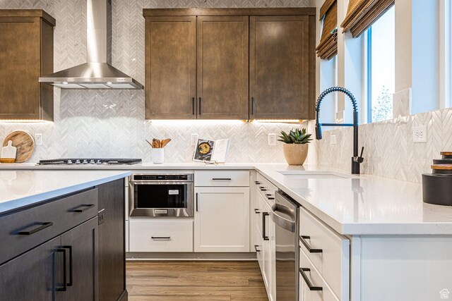 kitchen with wall chimney range hood, a sink, stainless steel appliances, wood finished floors, and white cabinetry