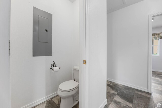 bathroom featuring electric panel, toilet, baseboards, and stone finish floors