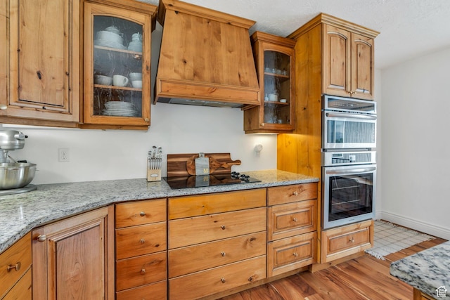 kitchen featuring custom exhaust hood, light stone counters, black electric stovetop, and glass insert cabinets