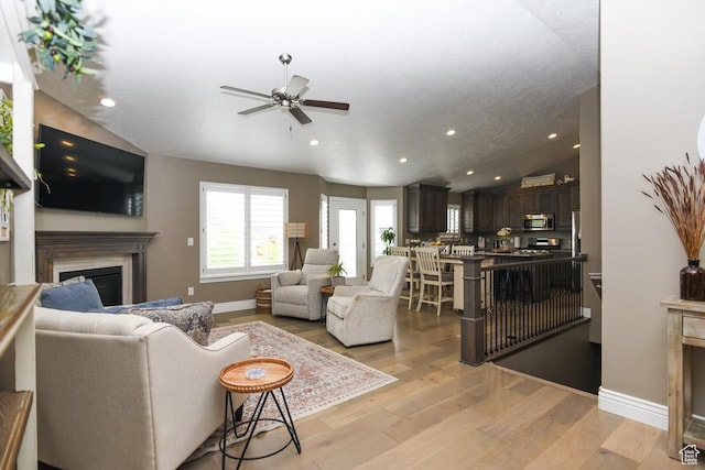living area with a glass covered fireplace, a ceiling fan, light wood-style flooring, vaulted ceiling, and recessed lighting