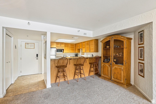 kitchen featuring stainless steel microwave, freestanding refrigerator, light colored carpet, brown cabinets, and a breakfast bar