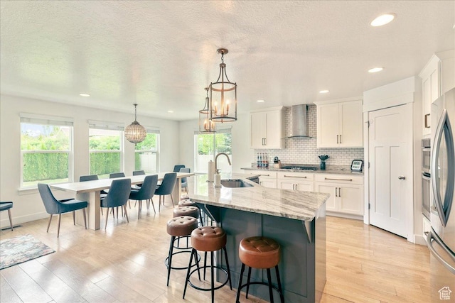kitchen featuring a sink, wall chimney exhaust hood, light wood-style flooring, white cabinetry, and a textured ceiling