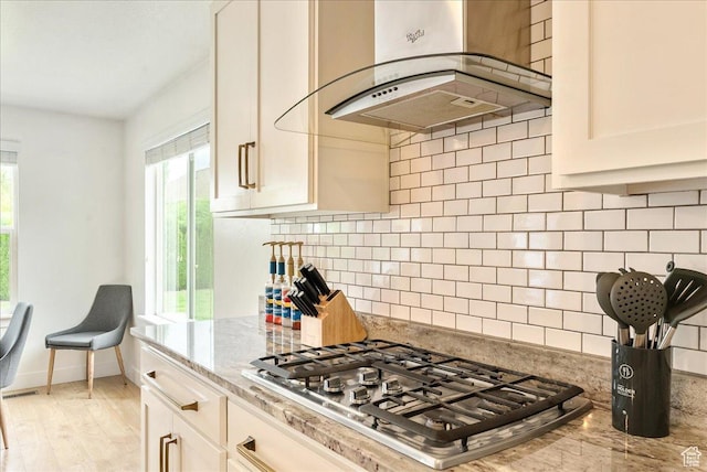 kitchen featuring stainless steel gas cooktop, range hood, light stone counters, decorative backsplash, and light wood-style floors
