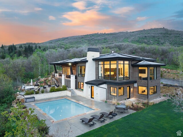 back of property at dusk with stairs, a balcony, an outdoor pool, a forest view, and a patio area