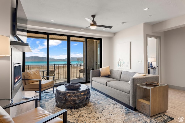 living area with a ceiling fan, wood finished floors, a warm lit fireplace, a water view, and recessed lighting