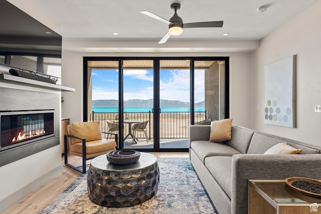 living area featuring light wood-style flooring, a glass covered fireplace, ceiling fan, a water view, and recessed lighting