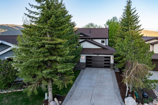 view of property hidden behind natural elements featuring driveway, stone siding, and roof with shingles