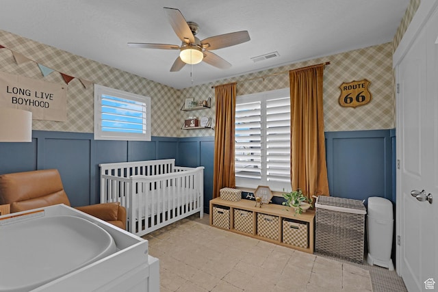 12473 S Desert Mesa Dr, Herriman, UT 84096 - bedroom featuring wallpapered walls, wainscoting, ceiling fan, a crib, and a decorative wall