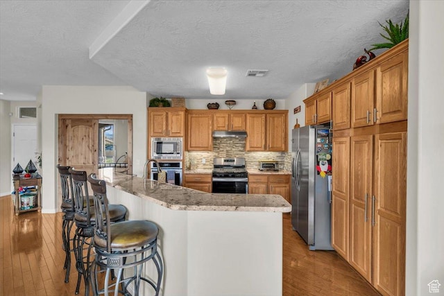 1646 W Sunkissed Dr, St George, UT 84790 - kitchen with appliances with stainless steel finishes, under cabinet range hood, a breakfast bar, light wood finished floors, and light stone countertops