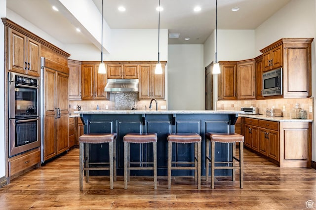 285 N Kings Peak Ct, Heber City, UT 84032 - kitchen featuring under cabinet range hood, built in appliances, brown cabinetry, a center island with sink, and backsplash