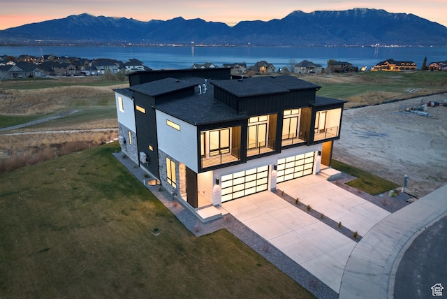 78 Watson Dr, Saratoga Springs, UT 84045 - back of house at dusk with a balcony, an attached garage, a shingled roof, concrete driveway, and a yard