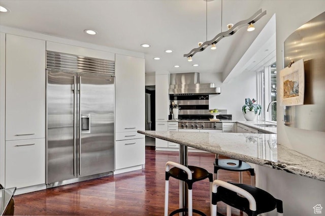 1074 E Raymond Rd, Fruit Heights, UT 84037 - kitchen with stainless steel built in fridge, wall chimney range hood, light stone counters, dark wood finished floors, and white cabinetry