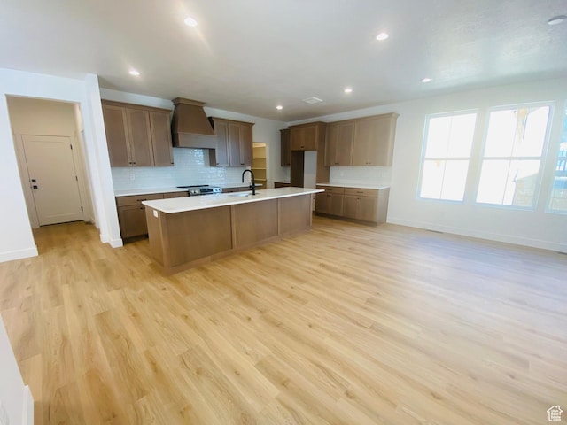 6511 W Lake Ave, South Jordan, UT 84009 - kitchen with custom exhaust hood, backsplash, light wood-style floors, light countertops, and a center island with sink