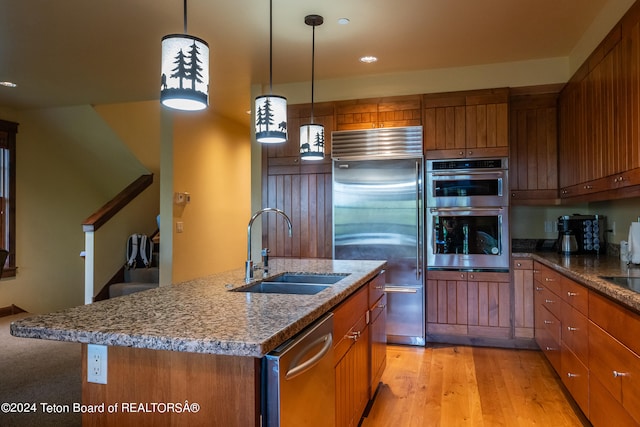 kitchen with appliances with stainless steel finishes, sink, decorative light fixtures, a kitchen island with sink, and light wood-type flooring