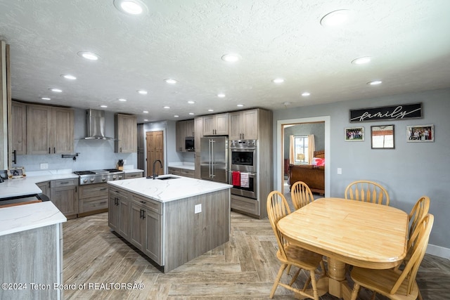 kitchen with appliances with stainless steel finishes, sink, a kitchen island with sink, light stone countertops, and wall chimney range hood