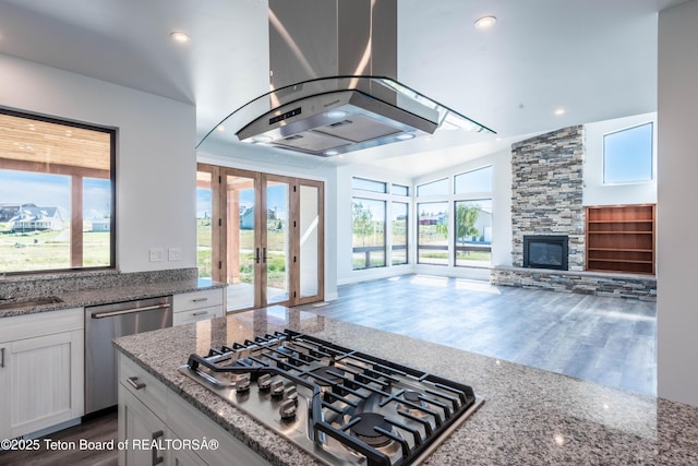 kitchen with appliances with stainless steel finishes, island range hood, a fireplace, light stone countertops, and white cabinets