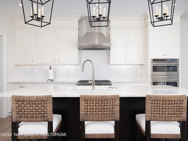 kitchen with stainless steel double oven, white cabinets, an island with sink, decorative backsplash, and hanging light fixtures