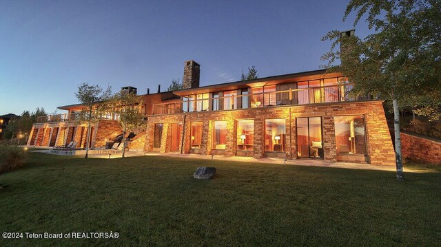 back house at dusk featuring a patio, a balcony, and a lawn