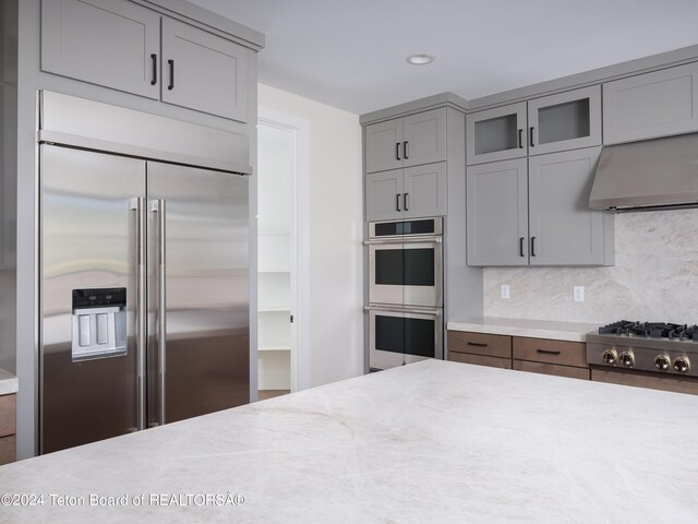 kitchen featuring gray cabinetry, stainless steel appliances, backsplash, ventilation hood, and light stone counters