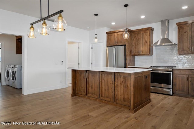 kitchen with appliances with stainless steel finishes, decorative light fixtures, a center island, washer and clothes dryer, and wall chimney exhaust hood