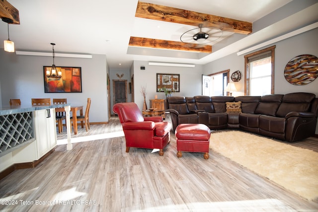 living room featuring ceiling fan with notable chandelier, light wood-type flooring, beamed ceiling, and a raised ceiling