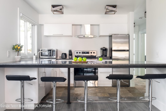kitchen featuring wall chimney range hood, white cabinets, appliances with stainless steel finishes, and a breakfast bar area