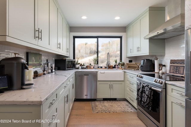 kitchen featuring light stone countertops, wall chimney exhaust hood, stainless steel appliances, tasteful backsplash, and light hardwood / wood-style flooring