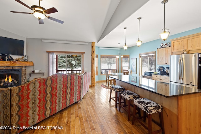 kitchen featuring a stone fireplace, light hardwood / wood-style flooring, dark stone countertops, stainless steel fridge, and vaulted ceiling