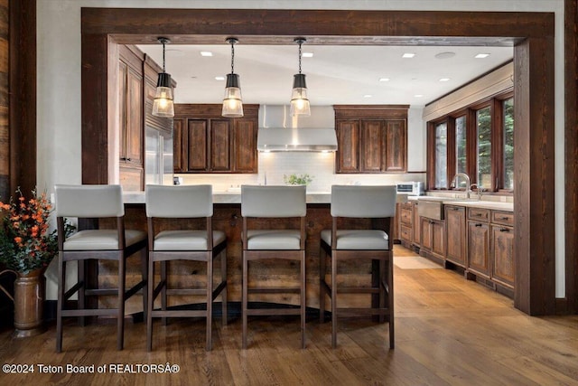 bar featuring decorative backsplash, wall chimney exhaust hood, sink, hardwood / wood-style flooring, and hanging light fixtures