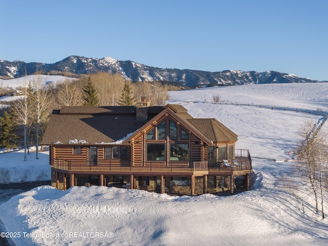 snow covered rear of property featuring a deck with mountain view, log exterior, and a chimney