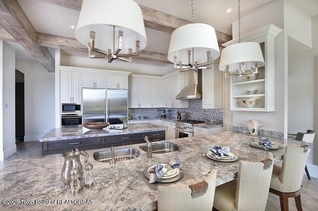 kitchen with pendant lighting, beamed ceiling, white cabinetry, built in appliances, and wall chimney range hood