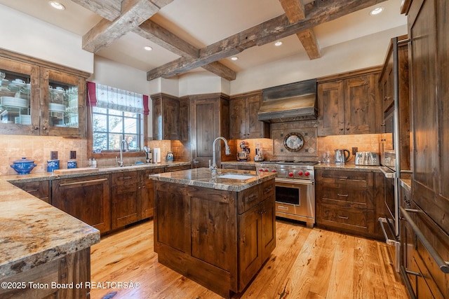 kitchen featuring an island with sink, backsplash, designer range, custom exhaust hood, and light hardwood / wood-style flooring