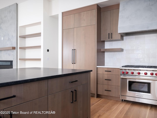 kitchen with wall chimney range hood, backsplash, light hardwood / wood-style floors, and designer stove