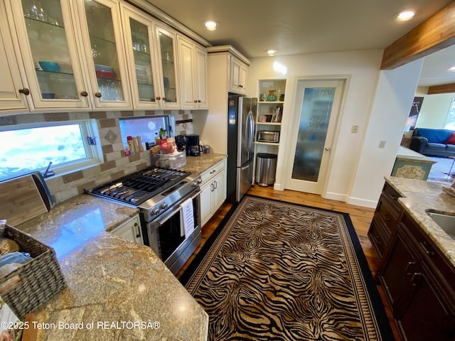 kitchen featuring light stone countertops, wood finished floors, appliances with stainless steel finishes, decorative backsplash, and glass insert cabinets