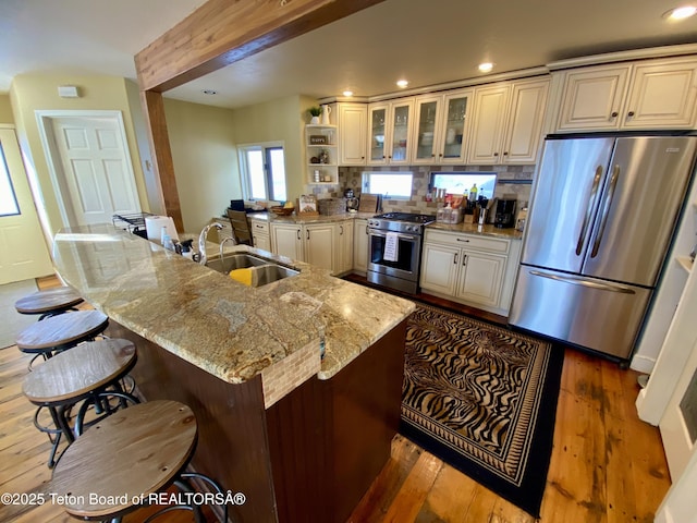 kitchen with stainless steel appliances, glass insert cabinets, a sink, an island with sink, and light stone countertops