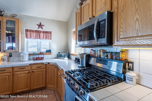 kitchen featuring tile countertops, light tile patterned floors, stainless steel appliances, vaulted ceiling, and glass insert cabinets