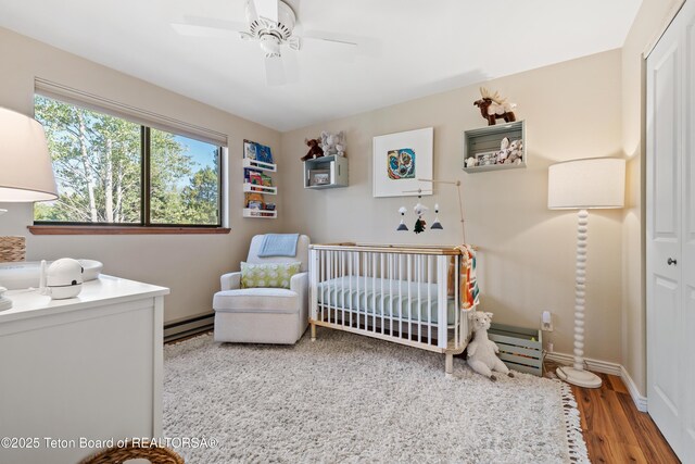 bedroom with wood finished floors, baseboards, a baseboard radiator, ceiling fan, and a nursery area